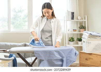 Happy asian young woman holding electric steam, hot iron press pile stripe shirt clothes on ironing board, housework after hygiene laundry at home. Housekeeping lifestyle, household of chores concept. - Powered by Shutterstock