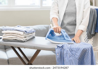 Happy asian young woman holding electric steam, hot iron press pile stripe shirt clothes on ironing board, housework after hygiene laundry at home. Housekeeping lifestyle, household of chores concept. - Powered by Shutterstock