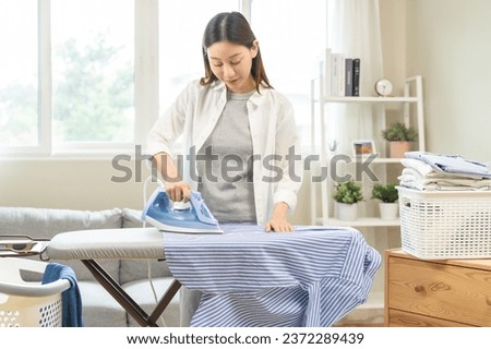 Happy asian young woman holding electric steam, hot iron press pile stripe shirt clothes on ironing board, housework after hygiene laundry at home. Housekeeping lifestyle, household of chores concept.