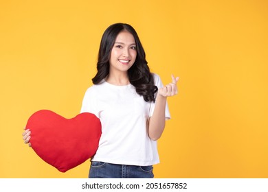 Happy Asian Young Woman Long Curly Hair Holding Red Heart Smiling Cute And Adorable Isolated On Yellow Background. Love And Valentines Day.