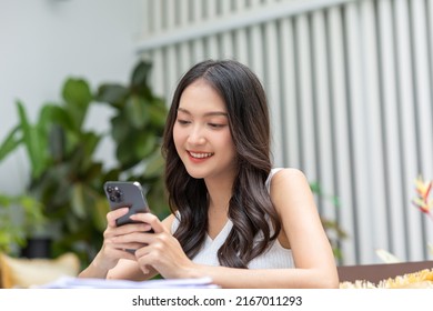 Happy asian young woman smile using smartphone surfing internet and enjoy with social media or online shopping at home. Modern wellness life of cute female looking at mobile phone fun and happiness - Shutterstock ID 2167011293