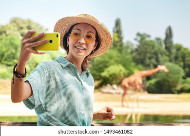 Happy Asian Zoology Student Girl Taking Selfie Photo On Smartphone While Giraffe Drinking From Lake