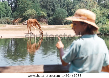 Happy asian zoology student girl looking at giraffe drinking from lake