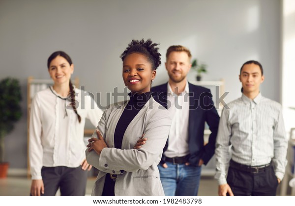 Happy aspiring black business woman in suit\
smiling at camera standing arms folded with team of workers behind.\
Portrait of ambitious female leader, executive, bank staff manager.\
Blurred background