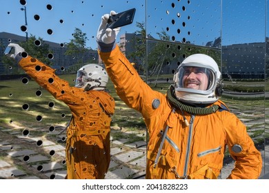 Happy astronaut wearing an orange spacesuit and space helmet holding mobile phone and taking selfie outdoors. Cheerful cosmonaut making a photo of himself standing against futuristic mirror wall