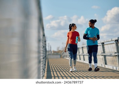 Happy athlete and her African American friend drinking water and communicating while walking over the bridge outdoors.  - Powered by Shutterstock