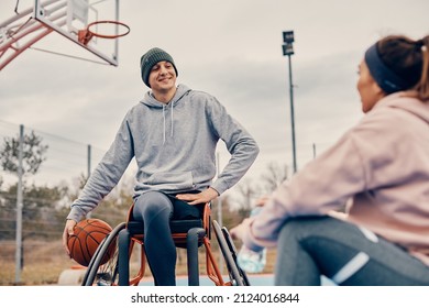 Happy Athlete In Wheelchair Communicating With Female Friend While Relaxing After Playing Basketball On Outdoor Sports Court.  