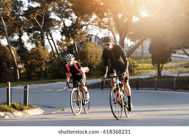 Happy Athletic Couple Enjoying Morning Ride On Racing Bicycles, Speeding On Desert Street. Young European Man And Woman Cyclists Wearing Stylish Sports Clothing Riding Road Bikes Outdoors In City