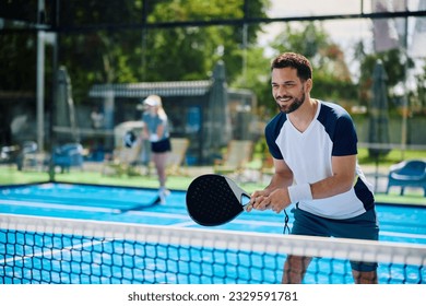 Happy athletic man standing by the net while playing padel with friends on outdoor tennis court.  - Powered by Shutterstock