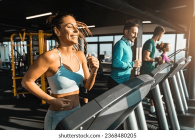 Happy athletic people jogging on treadmills in a health club. Beautiful people bathed in late afternoon sun running in a fancy gym. Selective focus of smiling slim fit woman exercising with friends. - Powered by Shutterstock