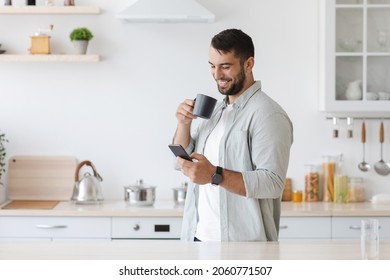 Happy attractive adult caucasian man drinking coffee and reading news at smartphone, chatting in social networks at kitchen interior. Guy enjoying morning tea, using online app, great offer and ad - Powered by Shutterstock