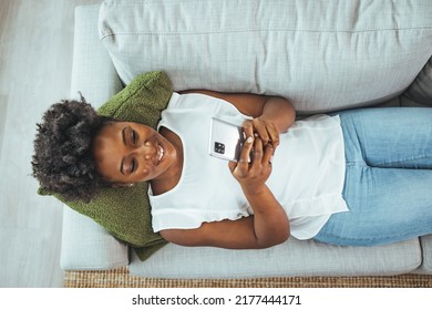 Happy attractive african american girl texting on a mobile phone. Cropped shot of an attractive young woman lying down on her sofa and using her cellphone at home. The internet holds  entertainment - Powered by Shutterstock