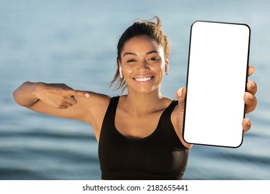 Happy Attractive Athletic Millennial Black Woman Pointing At Brand New Smartphone With White Empty Screen And Smiling, Recommending Newest Mobile Fitness App, Mockup, Seaside Background