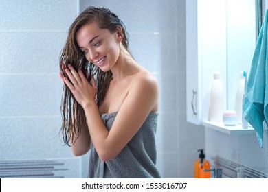 Happy Attractive Caucasian Joyful Brunette Woman In Bath Towel Applies Hair Conditioner In Bathroom After Shower At Home. Hair Care 