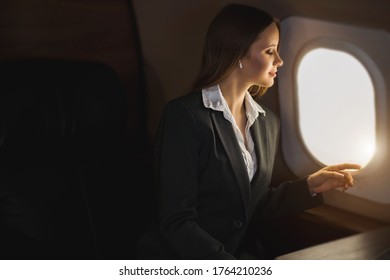 Happy attractive caucasian young businesswoman listening music using wireless earphones, looking at airplane window, admiring beauty of sky, enjoying flight in private jet.
