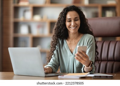 Happy attractive curly middle eastern young woman sitting at workdesk with laptop and notepads on, using smartphone and smiling, using newest business mobile app, office interior, copy space