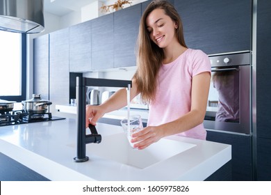 Happy Attractive Joyful Brunette Woman Pours Fresh Clean Filtered Purified Water For Drinking From A Faucet Into A Glass At Kitchen At Home. Healthy Lifestyle 