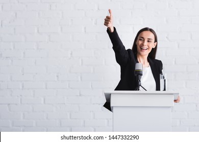 happy, attractive lecturer showing thumb up while standing on podium tribune