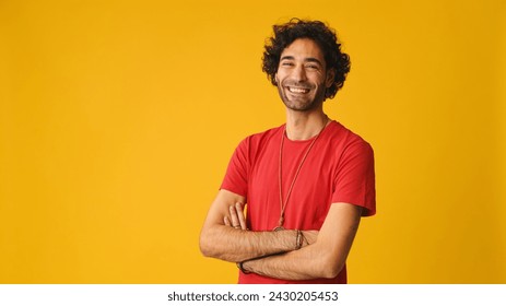 Happy attractive man with curly hair, dressed in red T-shirt,  crossing his arms and looking at camera isolated on yellow background in studio - Powered by Shutterstock