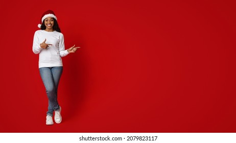 Happy attractive millennial black woman in Santa hat and casual outfit posing over red studio background, pointing at copy space and smiling, panorama, full length shot. Christmas deal concept - Powered by Shutterstock