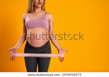 happy attractive pregnant woman in sports clothes does exercises with an elastic sports elastic band on yellow background, studio. health during pregnancy. sports at home. Motherhood, active pregnancy