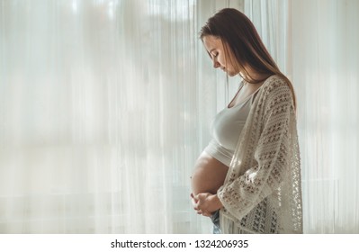 Happy Attractive pregnant woman standing near the window and holding her belly. Concepts of pregnancy and family. Motherhood. Pregnant belly. Awaiting baby
