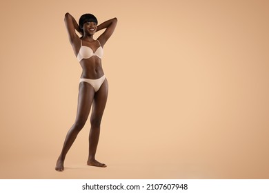 Happy attractive slender millennial black lady with hands up posing in nude underwear over beige studio background, showing her beautiful curves, panorama with copy space, full length shot