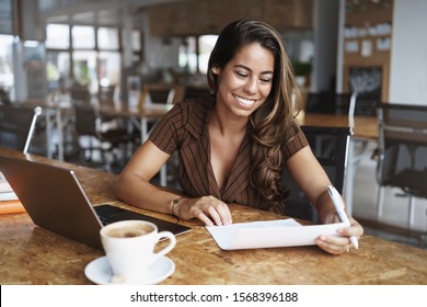 Happy attractive smiling latino woman sitting cafe, co-working urban space, grining delighted, reading paper and make corrections, prepare for office meeting, sit near opened laptop drink cappuccino