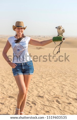 Happy attractive smiling woman wearing straw hat with eagle sitting on a hand in desert sand. Travel safari on vacation, sunny summer day. United Arab Emirates
