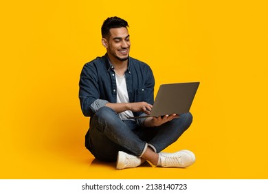 Happy attractive stylish young middle eastern guy sitting on floor and using modern notebook, typing on laptop keyboard and smiling, chatting with friends, isolated on yellow, copy space - Powered by Shutterstock