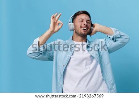Happy attractive tanned handsome man in casual basic t-shirt headphones listen cool sound show gesture Okay posing isolated on blue studio background. Copy space Banner Mockup. Music concept