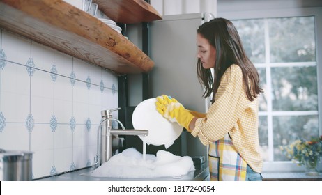 Happy attractive woman in apron and gloves washing dishes at home kitchen. Side view of pretty young housewife doing household. Beautiful female wash plates - Shutterstock ID 1817240054
