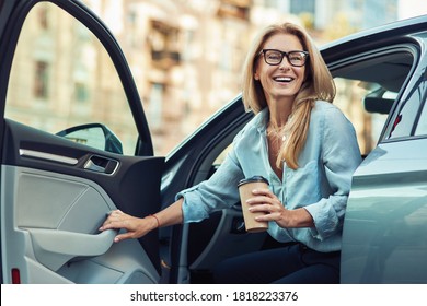 Happy attractive woman or business lady wearing eyeglasses holding cup of coffee and getting out of her modern car - Shutterstock ID 1818223376