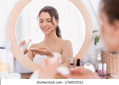Happy Attractive Woman Using Face Toner After Shower At Home, Applying Product On Cotton Pad In Front Of Mirror