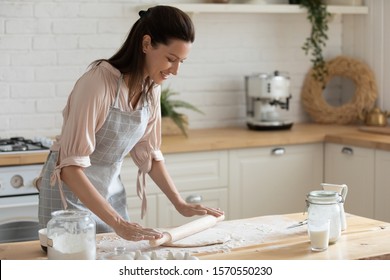 Happy attractive young adult woman lady housewife baker wear apron holding pin rolling dough on kitchen table baking pastry concept cooking cake biscuit doing bakery making homemade pizza at home