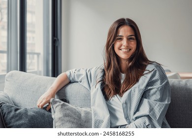 Happy attractive young adult woman girl sitting on sofa looking at camera in modern cozy home, smiling single 20s lady beautiful face relaxing on couch alone enjoy wellbeing posing for portrait - Powered by Shutterstock