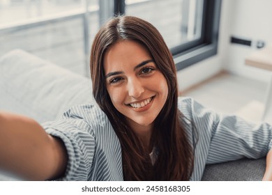 Happy attractive young adult woman housewife sitting on sofa looking at camera in modern cozy home, smiling single 20s lady beautiful face taking a selfie alone enjoy wellbeing posing for portrait - Powered by Shutterstock