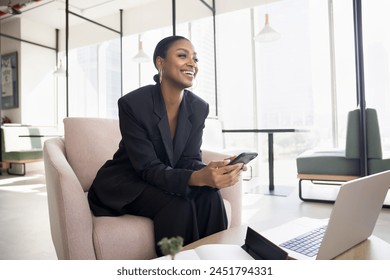 Happy attractive young African American business lady using Internet technology for job in co-working space, sitting in armchair at laptop, holding mobile phone, looking away, smiling - Powered by Shutterstock