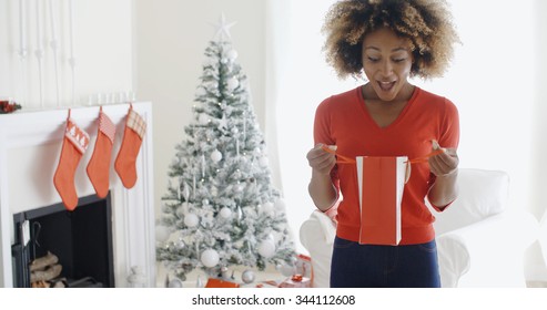 Happy Attractive Young African Woman Opening Christmas Gifts In Front Of The Tree Looking Into A Gift Bag In Surprise