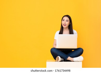 Happy Attractive Young Asian Woman With Laptop Computer Sitting Cross Legged And Looking At Empty Space On Yellow Studio Background