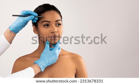 Happy attractive young black woman attending aesthetic clinic, unrecognizable doctor plastic surgeon making pre surgery marks on pretty lady face before beauty procedure, copy space, web-banner