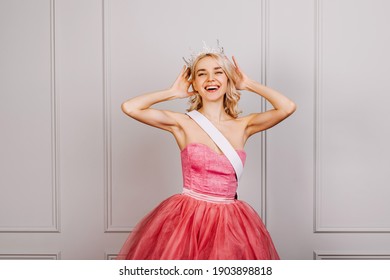 Happy Attractive Young Blonde Woman Wearing A Crown, Pink Dress And A White Ribbon, Smiling. Beauty Contest Winner Concept.