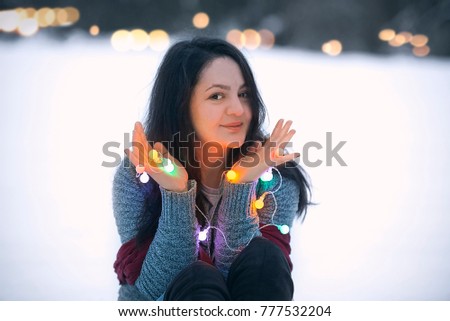 Happy attractive young brunette woman sitting in the snow, holding in her hands colorful lit string lights and playing with them.