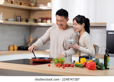 Happy Attractive Young Chinese Lady Supporting Her Husband While He Is Cooking Healthy Dinner For Family, Woman Drinking Coffee And Embracing Her Boyfriend Cheef, Copy Space