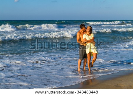 Happy attractive young couple in beachwear, enjoying a summer dusk at the beach, having fun walking barefoot, getting wet, kissing and teasing one another.