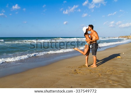 Happy attractive young couple in bikini and shorts enjoying summer dusk at the beach, practicing fitness exercises, having fun walking barefoot, kissing and teasing one another.