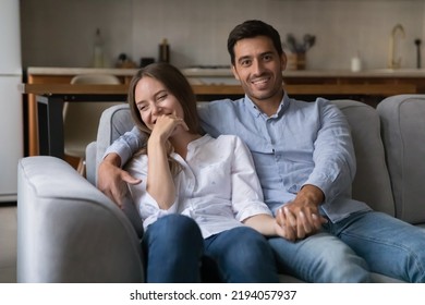 Happy Attractive Young Couple Home Head Shot Portrait. Millennial Husband And Wife Resting And Hugging On Couch, Looking At Camera, Smiling, Laughing, Talking On Video Call. Love, Relationship