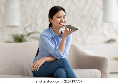 Happy attractive young japanese lady in casual outfit sitting on couch in living room, recording voicemail on mobile phone, communicating with friends via mobile app, looking at copy space