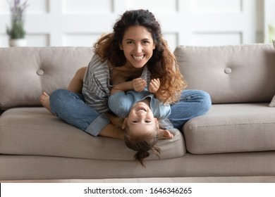 Happy Attractive Young Mom Holding Lying Upside Down Little Preschool Child Daughter Playing, Looking At Camera. Playful Laughing Joyful Two Generations Family Having Fun, Tickling On Sofa At Home.