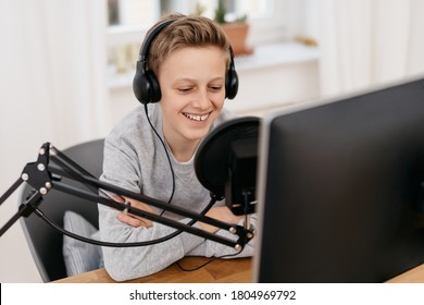 Happy attractive young teenage boy recording a podcast on a professional mike sitting with headphones in front of the computer - Powered by Shutterstock